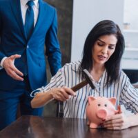 Worried woman holding hammer above piggy bank at table near collector with outstretched hand in room