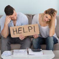 Worried Couple Holding Help Sign While Calculating Bills Worried Couple Holding Help Sign While Calculating Bills