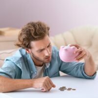 Young man tipping money from piggy bank at table