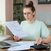 Young frustrated unhappy tired woman with financial troubles, sitting at kitchen table with papers, calculator and laptop computer, reading documents Young frustrated unhappy tired woman with financial troubles, sitting at kitchen table with papers, calculator and laptop computer, reading documents