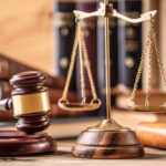 A judge’s gavel, a brass scale of justice, and legal books are arranged on a wooden table, symbolizing law and the judicial system.