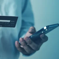 A person in a blue shirt holds a credit card and a smartphone, suggesting online payment or mobile banking. The mood is focused and modern.