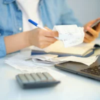 woman using a pen writing on bank account book while holding the bills to calculate in living room at home. Expenses, account, taxes, home budget concept