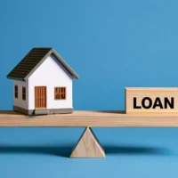 A model house sits on a wooden seesaw against a loan block. A concept for real estate finance and mortgage balancing.