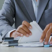 Businessman sorts papers at desk. Hands grip stacks of documents with focus. Office environment suggests planning or review. Scene conveys diligence and organization
